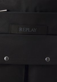Svart tygväska med en texturerad yta, med en framträdande "REPLAY" logotag och silverfärgade metalldragkedjespännen. Dragkedjestängning synlig.