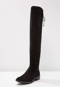 Bota negra de ante hasta la rodilla con tacón plano y textura suave. Cuenta con una punta redondeada y un pespunte decorativo a lo largo de la suela.