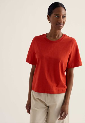 Žena s tmavými vlasmi, ktorá má na sebe zlaté kruhové náušnice, červené tričko s krátkym rukávom a béžové nohavice, stojaca pred jednoduchým svetlým pozadím.