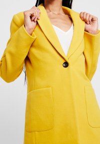 Manteau en mélange de laine jaune avec un col noté, fermeture à un bouton et deux poches plaquées à l'avant, avec une texture douce.