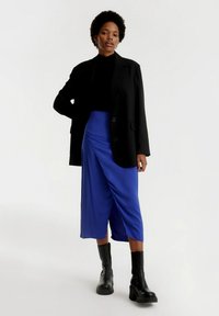 Zwarte oversized blazer over een getailleerde zwarte top, gecombineerd met een blauwe wikkelrok. Het model draagt zwart chunky hakken enkellaarsjes. Glad textuur overal.