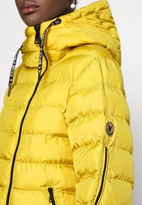 Chaqueta acolchada amarilla con capucha, con secciones acolchadas horizontales, cremallera negra y cordones, y un parche de logo redondo en la manga.