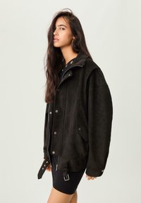 Jeune femme aux longs cheveux noirs portant des boucles d'oreilles créoles, une veste noire oversize avec des boutons en métal et une robe noire courte sur un fond uni.