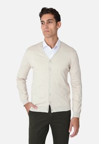 Cardigan in maglia beige con scollo a V e chiusura con bottoni, con maniche lunghe e orlo a coste, indossato sopra una camicia bianca con colletto.