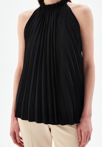 Svart plisserad halterneck-top med en volangkant, som visar upp ett texturerat tyg. Har en svävande design och bärs med beige byxor.