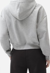 Felpa grigia cropped con cappuccio regolabile e orlo a costine, realizzata in tessuto morbido; indossata con pantaloni neri, con visione delle maniche e vista posteriore.