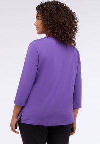 Nainen, jolla on kiharat hiukset, pukeutunut violettiin pitkähihaiseen puseroon ja mustiin housuihin, näkyy selkä kameraan, taustalla vaalea yksinkertainen tausta.