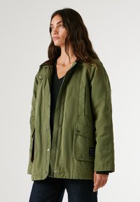 Olīvu zaļš džakets, kas izgatavots no kokvilnas auduma maisījuma, ar brūnu korduroja apkakli, spiedpogām un vairākām kabatām priekšpusē.