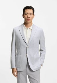 Blazer grigio chiaro con trama a spina di pesce, due tasche frontali, rever a lancia e chiusura con bottoni, abbinato a una polo color crema.