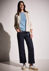 Ljus beige zip-up jacka, blå crew-neck tröja, mörka vida jeans och vita sneakers med gula sulor. Slät textur och avslappnad passform.
