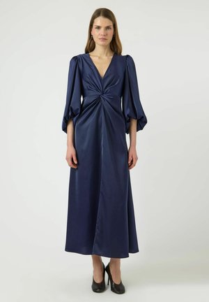 Kvinde i en marineblå satin midikjole med snoet detalje i taljen og puffede trekvartlange ærmer, iført sorte højhælede sko, stående.