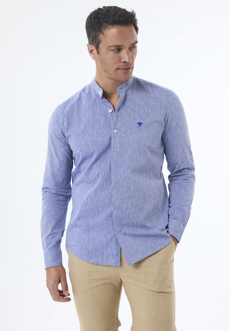 Camisa azul claro de manga larga con un acabado texturizado, cuello tipo mandarín y un pequeño logo azul marino en el pecho, combinada con pantalones cortos beige.