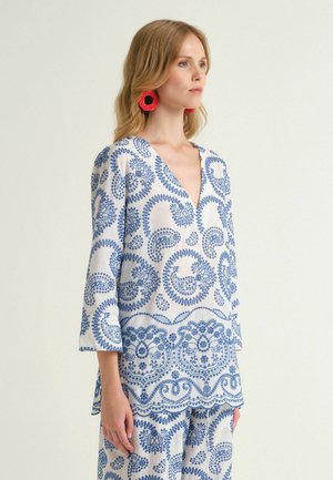 Donna con capelli biondi indossa una tunica bianca e pantaloni con motivi paisley blu e grandi orecchini rossi circolari, guarda di lato.