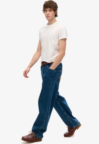 Camiseta de manga corta gris claro, vaqueros de denim azul holgados con un parche de logo y zapatos marrones con cordones y suela texturizada.