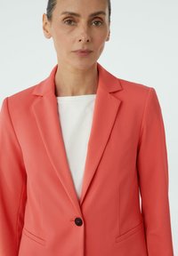 Koraal blazer van gladde stof, met een notch lapel, enkele knoopsluiting en zijzakken. Draagt over een witte top.