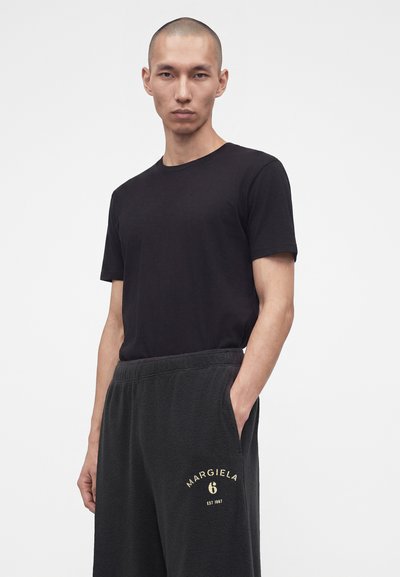Νέος άνδρας με κοντά μαλλιά, φοράει μαύρο T-shirt και σκουρόχρωμο φόρμα με το λογότυπο "Margiela 6" στο αριστερό μηρό, στέκεται μπροστά σε λευκό φόντο.