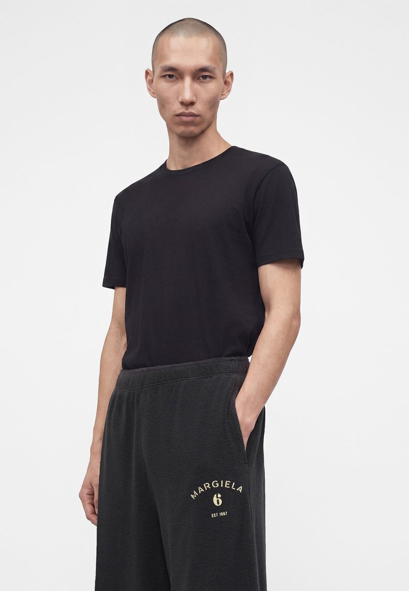 Junger Mann mit kurzem Haar, trägt ein schwarzes T-Shirt und dunkle Sweatpants mit "Margiela 6"-Logo am linken Oberschenkel, steht vor weißem Hintergrund.