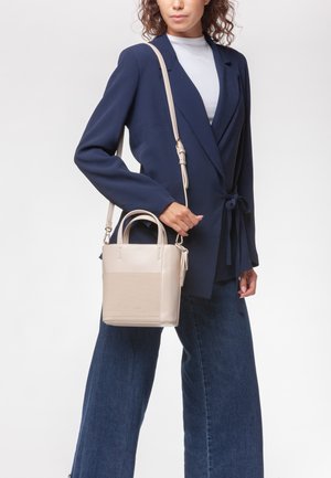 Borsa a mano in pelle beige con fondo testurizzato, doppi manici superiori e tracolla staccabile, indossata sopra un blazer blu navy.
