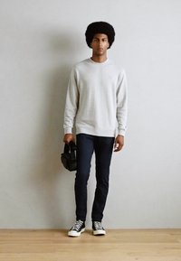 Light gray sweatshirt, dark blue skinny jeans, and black high-top sneakers. Model holds a black bag, standing against a plain wall.