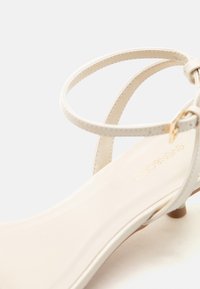 Κοντινό πλάνο μπεζ σανδαλιού με λεπτά λουράκια στον αστράγαλο και μικρό τακούνι, που εμφανίζει τις λεπτομέρειες των ραφών και την χρυσή αγκράφα.