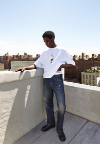 Witte T-shirt met korte mouwen en grafische print, blauwe spijkerbroek en zwarte schoenen. Staande op een balkon met een stadsgezicht op de achtergrond.