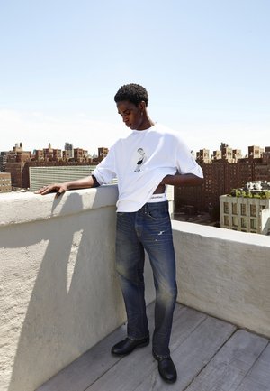 Witte T-shirt met korte mouwen en grafische print, blauwe spijkerbroek en zwarte schoenen. Staande op een balkon met een stadsgezicht op de achtergrond.