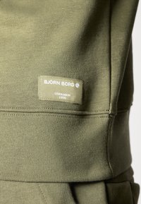 Felpa verde oliva con tessuto testurizzato, caratterizzata da un'etichetta rettangolare che riporta la scritta "BJÖRN BORG STOCKHOLM 1956" in bianco.