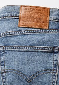 Jeans de denim azul claro con un parche de cuero que presenta texto y diseño en relieve, costuras visibles y detalles en los bolsillos traseros. Modelo 511, W 32 L 32.