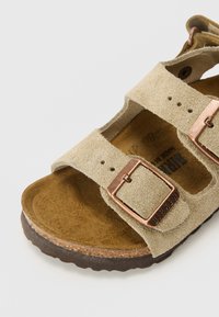 Beige suedetud sandaal, millel on kaks reguleeritavat rihma vasktooni pandlitega, tekstureeritud tallaga ja pruunist kummist välistald.