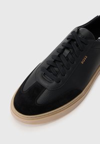 Zwarte leren en suède sneaker met een gestructureerde afwerking, ronde neus, vetersluiting en beige rubberen zool. Gouden "BOSS" logo aan de zijkant.