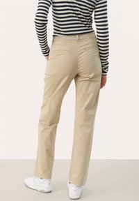 Osoba oblečená v béžových kalhotách, pruhovaném černobílém tričku s dlouhým rukávem a bílých teniskách, stojící s rukama v kapsách.