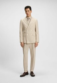 Beżowy dwurzędowy garnitur z podwójnymi guzikami, spodniami z płaskim przodem, klapami w kształcie notcha i dopasowanym krawatem. Brązowe buty wizytowe dopełniają całość.