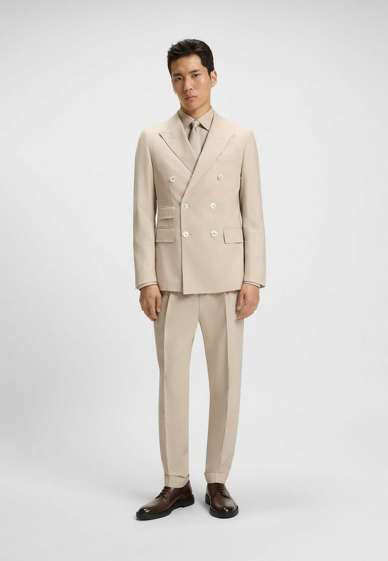 Beżowy dwurzędowy garnitur z podwójnymi guzikami, spodniami z płaskim przodem, klapami w kształcie notcha i dopasowanym krawatem. Brązowe buty wizytowe dopełniają całość.