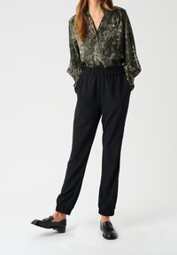 Femme portant un pantalon noir à poignets élastiques, des mocassins noirs et un chemisier à manches longues à motif vert, les mains dans les poches sur fond blanc.