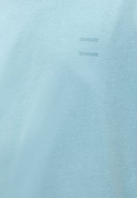 Ανοιχτό μπλε ύφασμα με δύο μικρά, διακριτικά λογότυπα "SAMSØE" εκτυπωμένα κάθετα σε ελαφρώς πιο σκούρα απόχρωση.