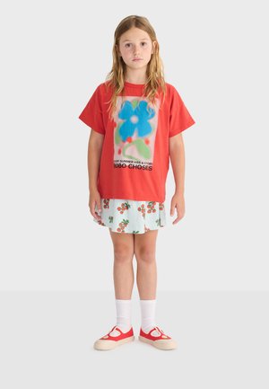 Young girl with long hair wearing a red oversized T-shirt with a blue flower print, white cherry-patterned skirt, white socks, and red shoes.