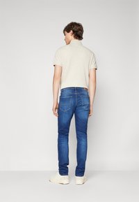 Egy férfi áll, háttal felénk fordulva, bézs színű, rövid ujjú pólót, kék farmernadrágot és fehér sportcipőt viselve, egy sima világos háttér előtt.