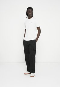 Balti marškinėliai su apvalia apykakle ir nedideliu logotipu ant krūtinės, derinami su laisvai krentančiomis juodomis krovimo kelnėmis su šoninėmis kišenėmis. Baltos sportbačiai.