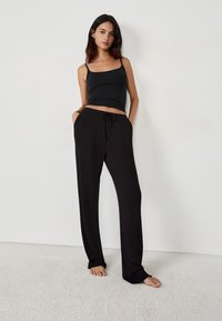 Czarny bezrękawnik i luźne czarne spodnie z kieszeniami. Miękka tkanina o swobodnym kroju i sznurkiem w talii. Na bosaka na teksturowanej podłodze.