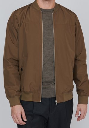 Mann trägt eine braune Bomberjacke mit Reißverschluss über einem olivgrünen Pullover und schwarzen Hosen, steht vor einfachem grauem Hintergrund.