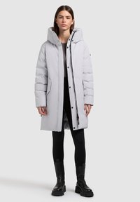 Gaiši pelēka polsterēta jaka ar augstu apkakli, priekšējo rāvējslēdzēju, spiedpogām un sānu kabatām. Kombinēts ar melnām legingām un masīvām melnām zābkām.