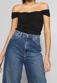 Musta olkapäiden päältä avattava paita, jossa on poimuttunut pinta, yhdistettynä korkeavyötäröisiin, löysiin sinisiin farkkuihin, joissa on klassinen nappi- ja vetoketjukiinnitys.