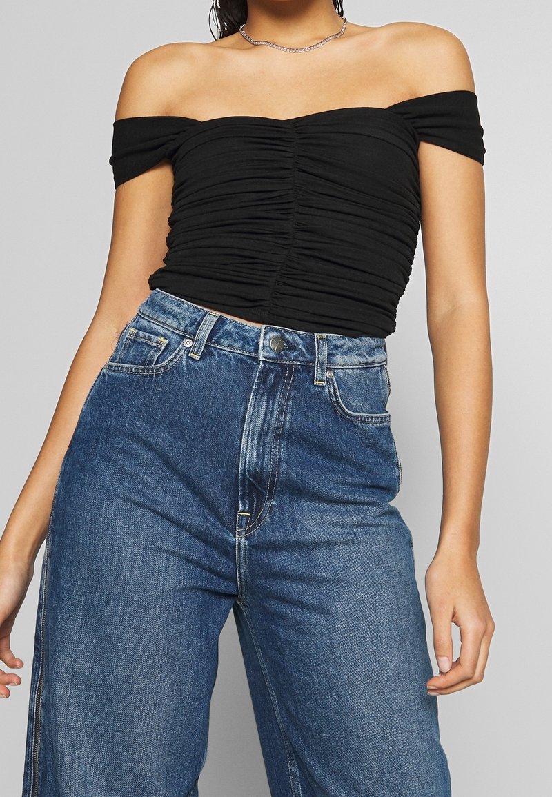 Musta olkapäiden päältä avattava paita, jossa on poimuttunut pinta, yhdistettynä korkeavyötäröisiin, löysiin sinisiin farkkuihin, joissa on klassinen nappi- ja vetoketjukiinnitys.