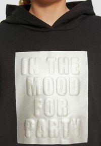 Zwarte hoodie met een gestructureerde witte graphic met "IN THE MOOD FOR PARTY" in vet, 3D-letters op de voorkant. Gezellig materiaal, casual ontwerp.