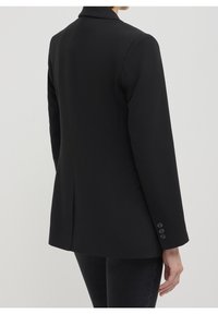 Femme portant un blazer noir ajusté avec des détails de boutons sur le poignet, vue de dos sur un fond gris clair.