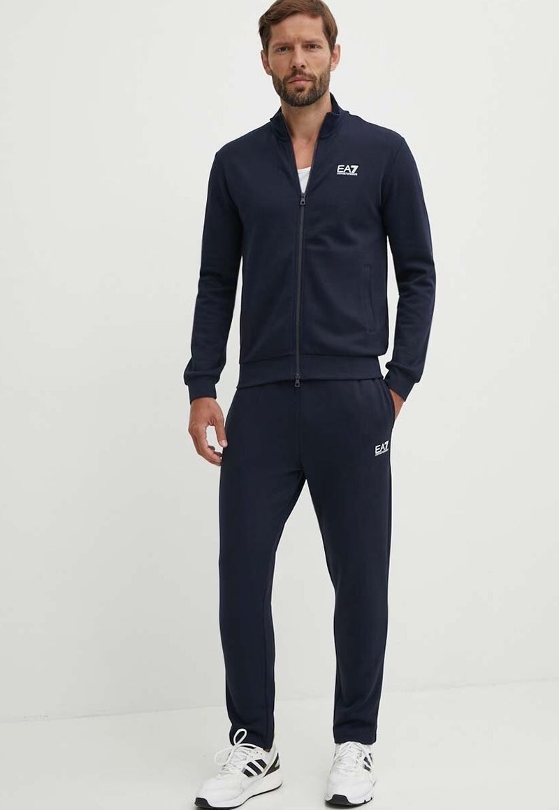 Completo tuta navy composto da giacca con zip e pantaloni con vita elastica, realizzato in tessuto morbido con design del logo sul petto e sulla gamba.