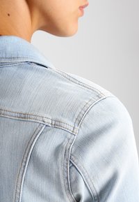 Hellblaue Jeansjacke mit klassischem Kragen, sichtbaren Nähten und weicher Textur. Fokus auf Schulter- und Halsbereich.