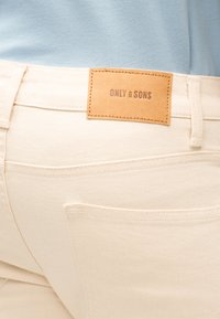 Μπεζ παντελόνι με ταμπέλα από δέρμα σε καφέ απόχρωση με την ένδειξη "ONLY & SONS", φορεμένο με γαλάζιο τοπ, που δείχνει τις ραφές και λεπτομέρειες στην πίσω τσέπη.