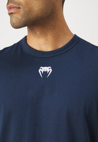 Mørkeblå t-skjorte med kort erme laget av bomull, med en hvit slange-logo på brystet og klassisk rund hals.