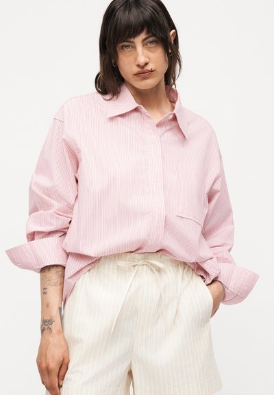 Άτομο με σκούρα μαλλιά μέχρι τους ώμους που φοράει άνετο ροζ ριγέ πουκάμισο και κρεμ ριγέ σορτς με κορδόνι, με τατουάζ στο αριστερό χέρι.